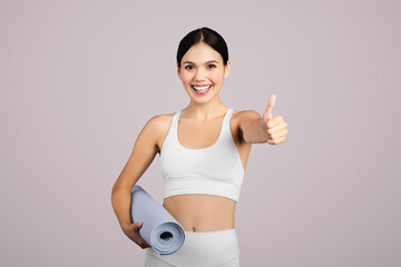 Enthusiastic fitness woman with yoga mat gives thumbs up over light background