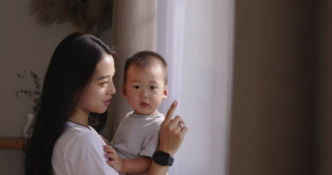 Asian beautiful mother and little child looking out window. Chinese, Japanese, Korean, Asian. Magical connection between mother and child, strong emotional attachment and adorable cute family scenes