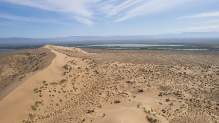 A large sand dune in the middle of the steppe. Drone view of a huge pile of sand. Tourists are walking, enjoying the view. A river runs in the distance and grass grows. The sky with white clouds