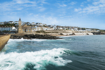 Waves along the sea wall in the foreground and the buildings of Porthleven, Cornwall lining the promenade and beach in the background.