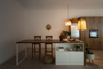 Hat, wicker basket and fresh bread on wooden island table in modern kitchen with glowing lamps