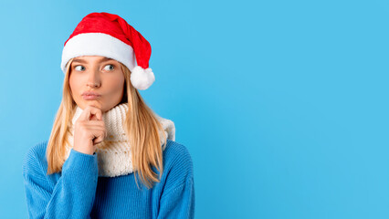 Pensive young woman in Santa hat, looking at free space on blue studio background, panorama