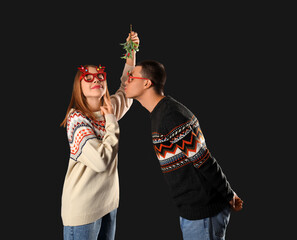 Young couple kissing under mistletoe branch on dark background
