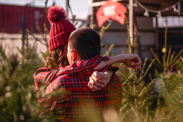 Couple in checkered red shirts, knitted hats hug, face to face among green Christmas tree market