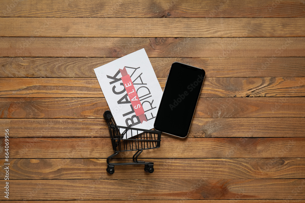 Sticker Black Friday sale greeting card, shopping cart and mobile phone on wooden table