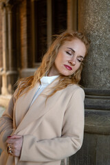Fototapeta premium Beautiful young woman with closed eyes in beige trench coat outside near column l. Vertical frame.