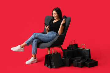 Young Asian woman with gift boxes, mobile phone, credit cards and shopping bags sitting on chair...