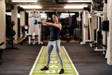 A sportswoman is standing in a gym and swinging with kettlebell.