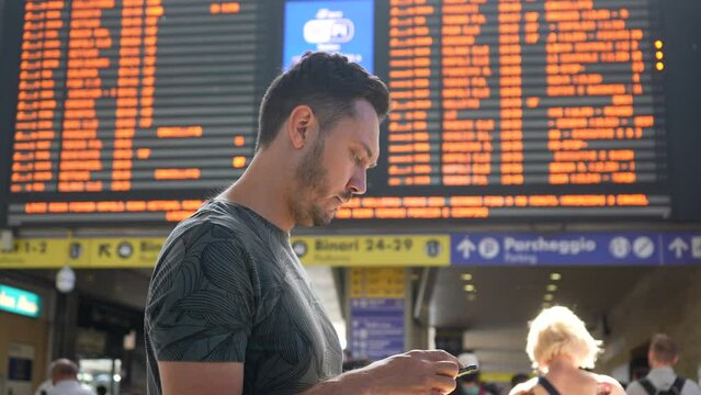 Bearded male tourist in t-shirt holding using mobile phone checking flight departure standing on blurred background of flight schedule board. Concept of vacations and travel. Shooting in slow motion.