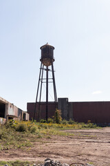 Water tower is set around an abandoned area. This rusty metal structure stands tall against a blue sky. Empty buildings all around showing signs of decay. Nature taking over in an apocalyptic scene.