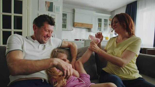 A positive family of american dream play, have fun and tickle each other in the room on the sofa. Mother, father and daughter laugh merrily and heartily. Little girl twitches her legs. The concept of