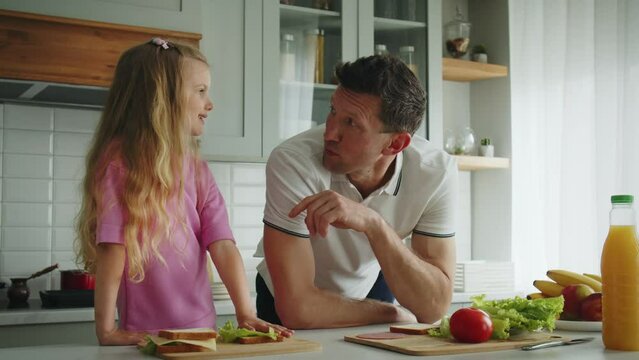 The smiling loving father and daughter standing in the kitchen at the table, he helps cooking sandwiches in playful mood and jokes with her. Showing index finger gesture. The concept of happy