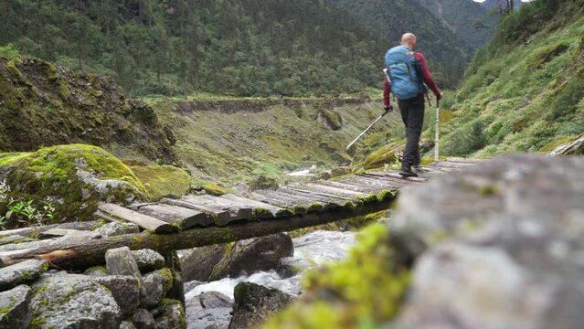 Man with backpack and trekking poles crossing mountain creek wooden bridge during Makalu Barun National Park trek in Nepal. Mountain hiking and active people concept image.