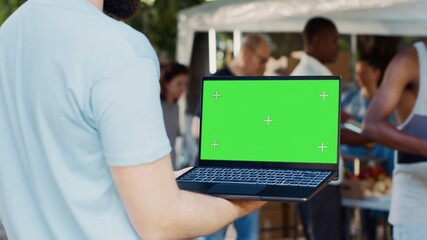 Caucasian man shown in a close-up holding digital laptop with isolated chromakey template. Male volunteer at an outdoor food bank grasping wireless computer displaying blank green screen. - obrazy, fototapety, plakaty
