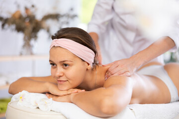 Positive young adult woman relaxing in cozy spa center while professional masseuse doing sports...