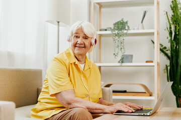 An elderly woman with headphones and a laptop sits on the couch at home and works.