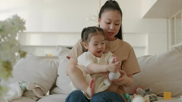 Medium shot of young Asian woman helping her little daughter to take off socks while sitting on couch in minimalist style apartment at daytime