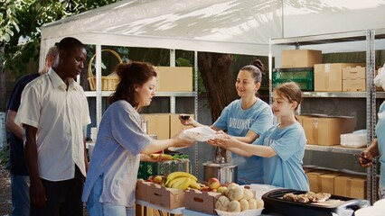 Caucasian girl packs free food to be distributed to the underprivileged with assistance from other charity workers. Female volunteers serving hot meals to the homeless and hungry at outdoor food bank. - obrazy, fototapety, plakaty