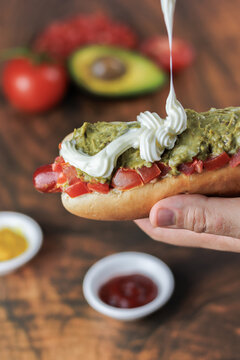 completo italiano: Close-up vertical. Adding mayonnaise, over tomato and vianesa on hot dog bun, held with one hand, tomato, palta and mustard on a wooden table. Typical Chilean food concept