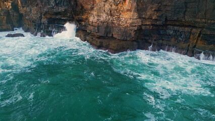 Naklejka premium Foamy waves washing rocky coastline in slow motion. Aerial dark coastal cliffs
