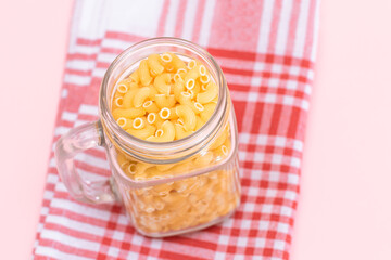 Uncooked Chifferi Rigati Pasta in Glass Jar on Checkered Red Towel on Pink Background. Fat and Unhealthy Food. Scattered Classic Dry Macaroni. Italian Culture and Cuisine. Raw Pasta
