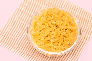 Uncooked Fusilli Pasta in Glass Jar on Bambo Mat on Pink Background. Fat and Unhealthy Food. Classic Dry Spiral Macaroni. Italian Culture and Cuisine. Raw Pasta