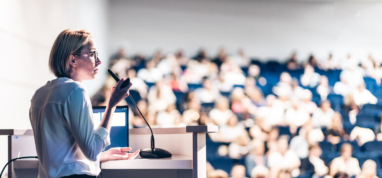 Female speaker giving a talk on corporate business conference. Unrecognizable people in audience at conference hall. Business and Entrepreneurship event