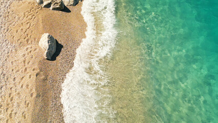 Vertical close aerial view of turquoise sea water, white wave, rock and little pebbles