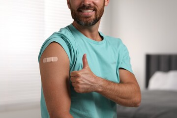 Man with sticking plaster on arm after vaccination showing thumbs up in bedroom, closeup - obrazy, fototapety, plakaty