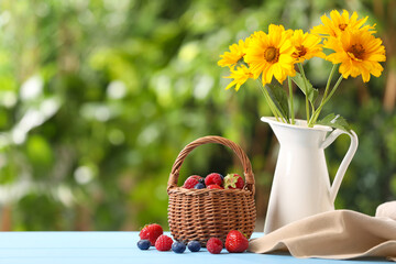 Fototapeta na wymiar Wicker basket with different fresh ripe berries and beautiful flowers on light blue table outdoors, space for text
