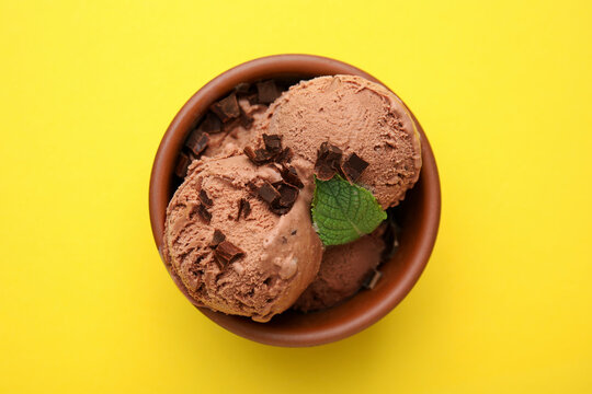Bowl of tasty ice cream with chocolate chunks and mint on yellow background, top view