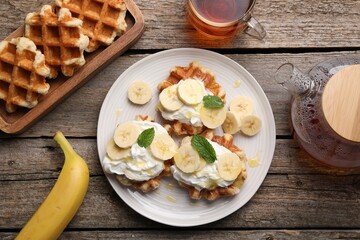 Delicious Belgian waffles with banana and whipped cream served on wooden table, flat lay