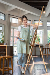 Portrait of young woman painting holding a palette and paintbrush in front of a canvas in a painting studio.