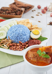 Nasi kerabu served with anchovies, sambal, peanuts, cucumbers and boiled egg. Accompanied by curry chicken rendang. Isolated against a white background with a background of spices. Selective focus.