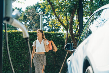 Young woman travel with EV electric car charging in green sustainable city outdoor garden in...