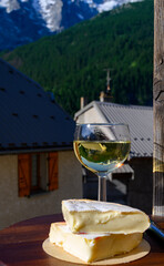 Reblochon French cheese made in Haute-Savoie, white Roussette de Savoie or Vin de Savoie wine with view on old alripne village and Hautes Alpes mountains snow tops in summer