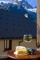 Reblochon French cheese made in Haute-Savoie, white Roussette de Savoie or Vin de Savoie wine with view on old alripne village and Hautes Alpes mountains snow tops in summer