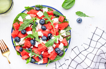 Juicy watermelon salad with feta cheese, blueberries and arugula on white table background, top view
