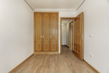 Fototapeta premium Empty bedroom with a built-in three-door oak wardrobe with handles, a skirting board of the same material, smooth white painted walls and plaster moldings on the ceiling