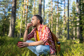 Man hiker backpacker drinking tea eating snack sitting in forest has halt, resting. Tourist guy in summer warm day in woodland. Camping hiking tourism concept. Outdoors activity recreation on nature.