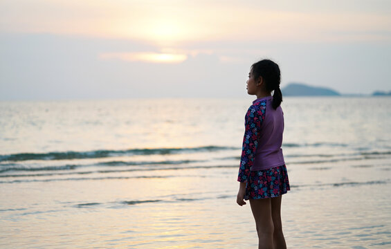 Portrait Asian child girl standing on the beach alone, view 45 degrees from behind, sunset light, beautiful calm sea and sky, landscape image with space for copy and design.