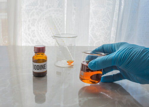 Gloved hand holding a beaker with Lugol's solution. Little glass jar with lugol on the table.