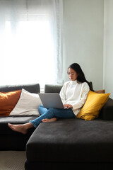 Young Asian woman working with laptop at home sitting on the couch in cozy apartment living room. Copy space.