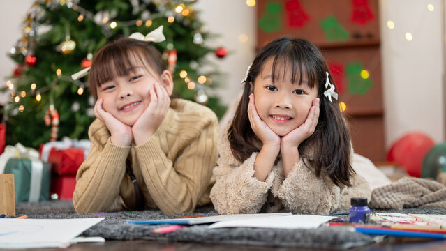 Two adorable and happy young Asian girls are lying on the floor near a Christmas tree together.