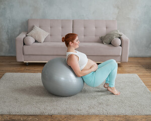 Happy pregnant woman lies with her back on a fitball and keeps her hands on her tummy at home.