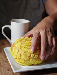 Woman's hand taking a piece of Mexican sweet bread from a white plate on a wooden table. Concept of hands handling food. Taking a yellow CONCHA with a cup of coffee on the side, vertical image