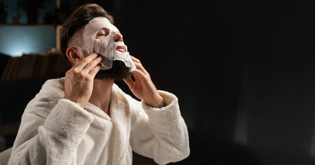 Bearded attractive man in bathrobe applying skincare facial face mask for anti wrinkle in his bedroom. Moisturizer facial sheet mask for men.