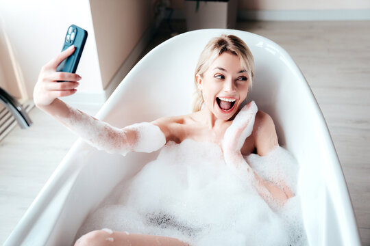 Young beautiful sexy woman having fun while lying in bathtub full of foam at home. Charming smiling model relaxing in luxury bath interior. Female holding smartphone, taking selfie photos, video call