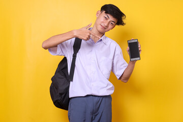 Young Asian student in schoolwear pointing at blank smartphone screen mockup for educational ads with happy expression isolated on yellow background