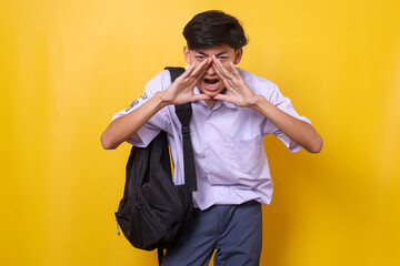 Excited young Asian male teen student wearing school uniform and backpack holds hand near mouth and...